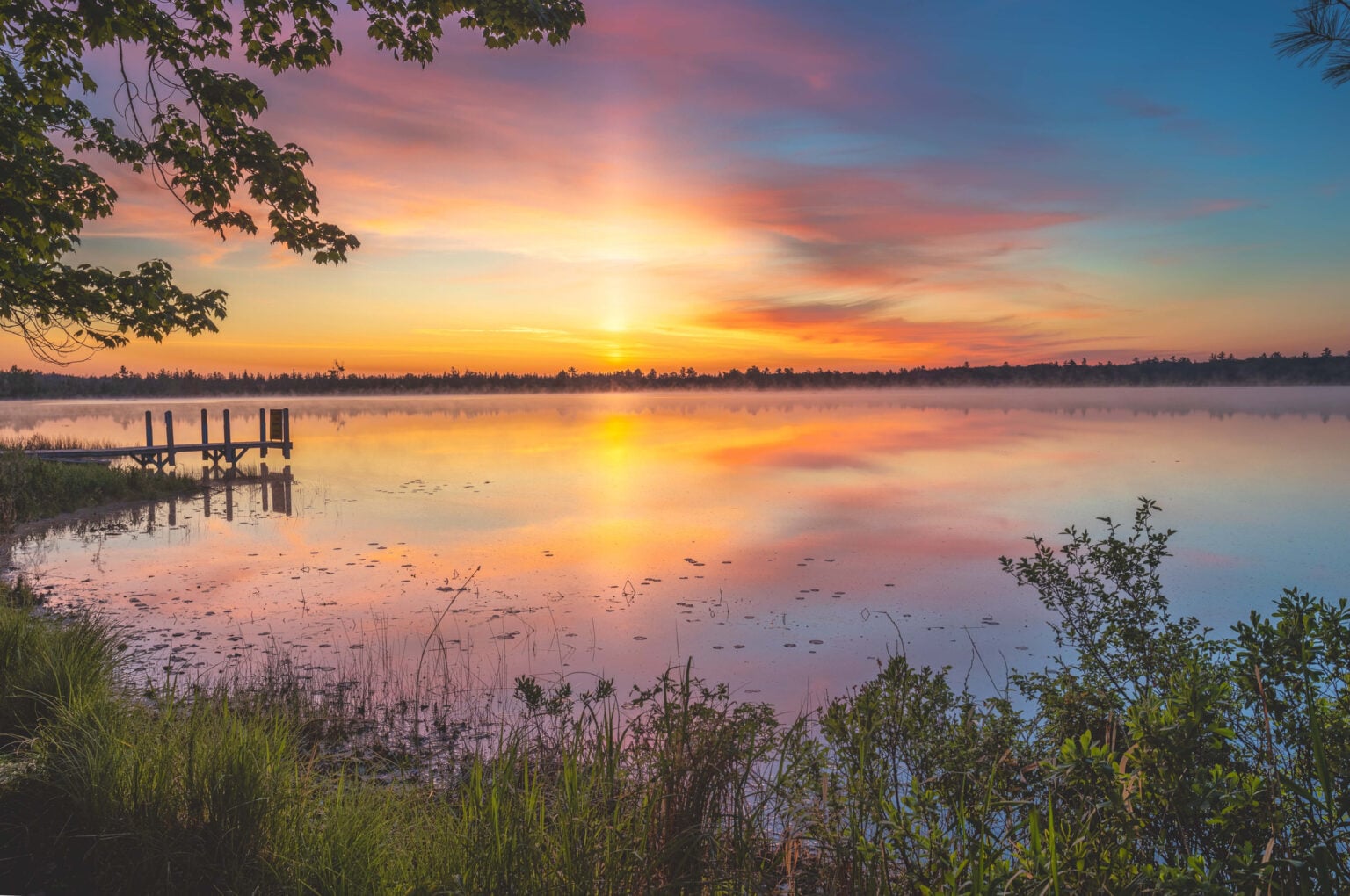 Sunset over a lake with a short pier and foliage showing on the edges.