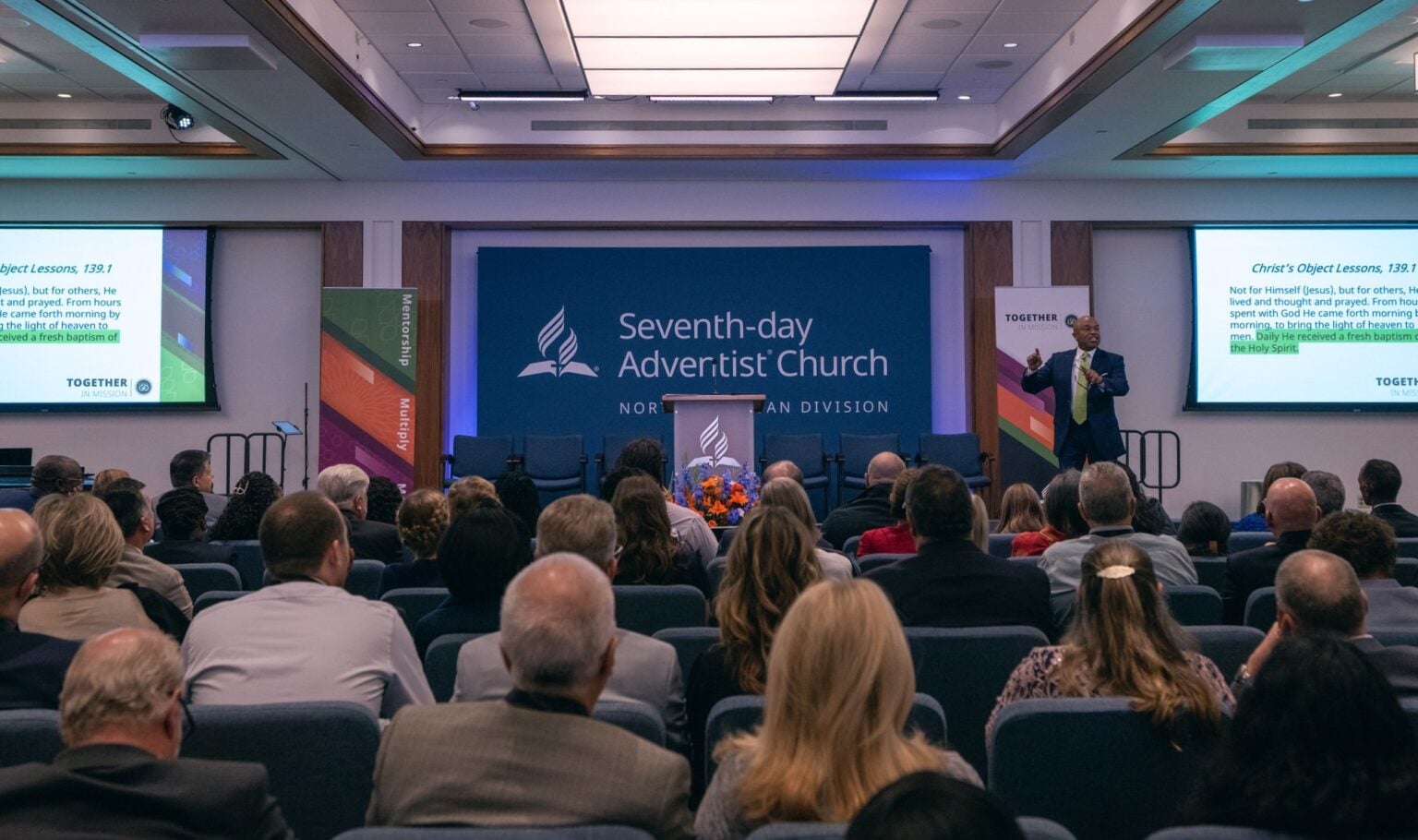 Photo of a large business meeting with a seated crowd. Taken from the back and towards the front with a presenter and screens.