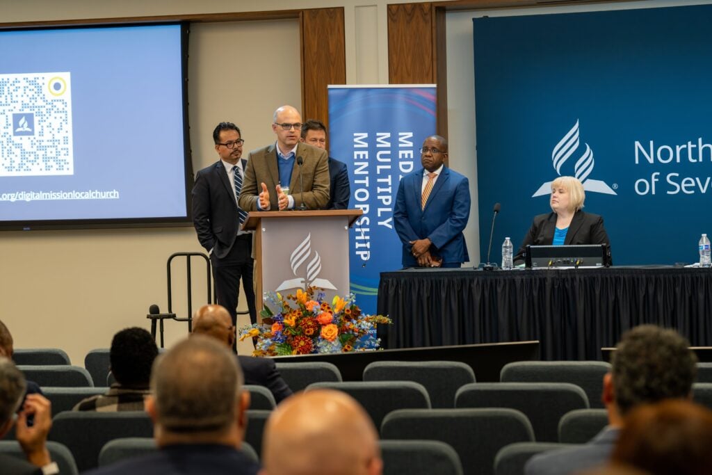 A man speaks from a podium while three other men stand nearby and a woman sits at a table beside him.