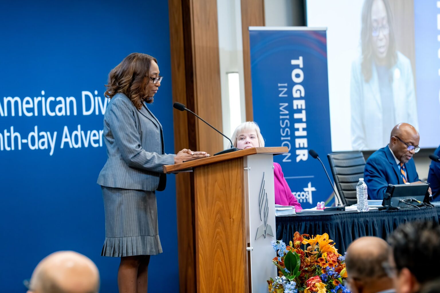African-American woman in business suit at a podium on stage addressing a professional meeting.