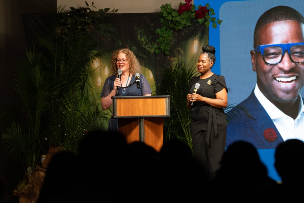Two women speak on stage at a gala in front of a photo of a smiling man.