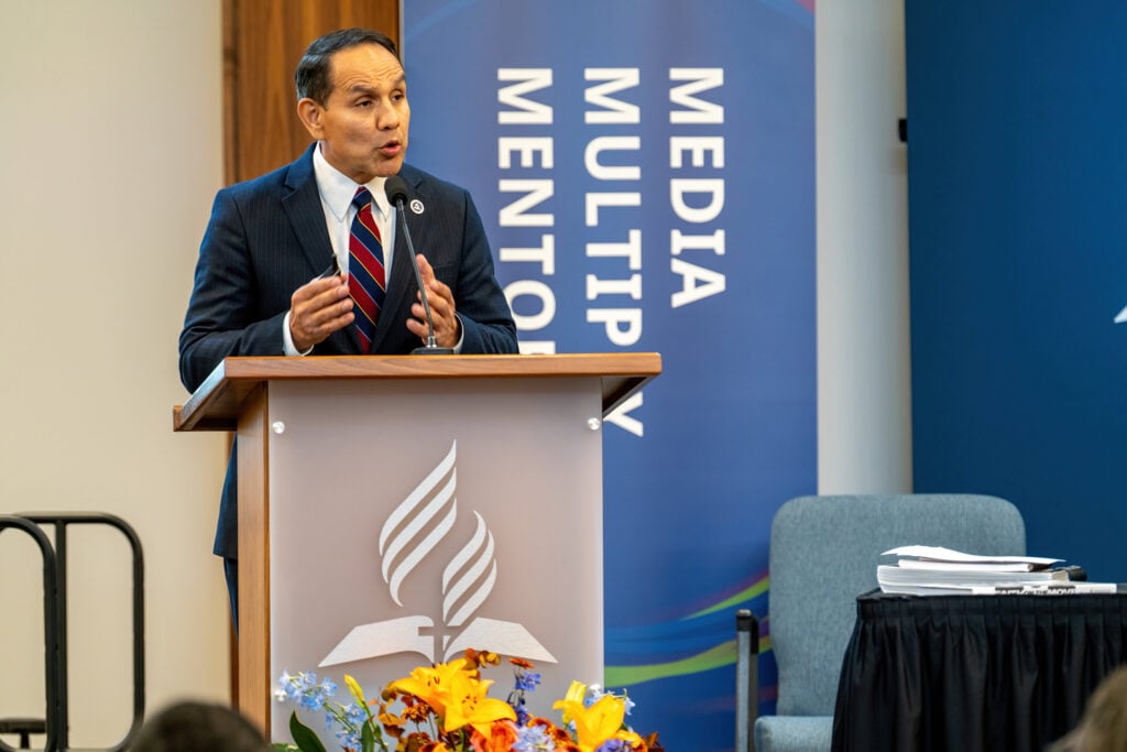 Man speaks at podium with media, multiply, mentorship banner behind it.