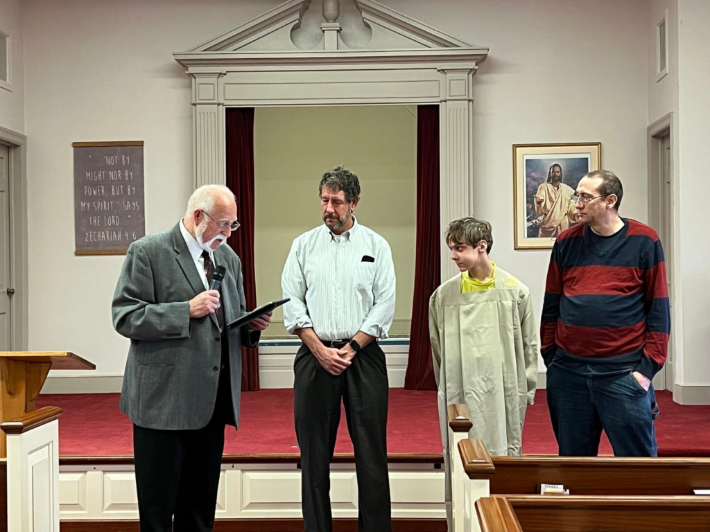 Four men stand in front of a congregation, one with a mic in hand, reading