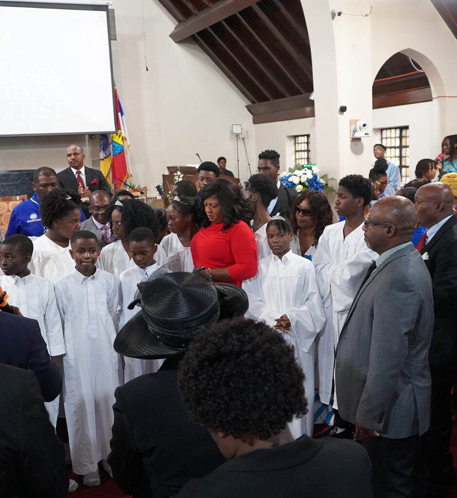 Young people are in white robes preparing for baptism, surrounded by adults. 