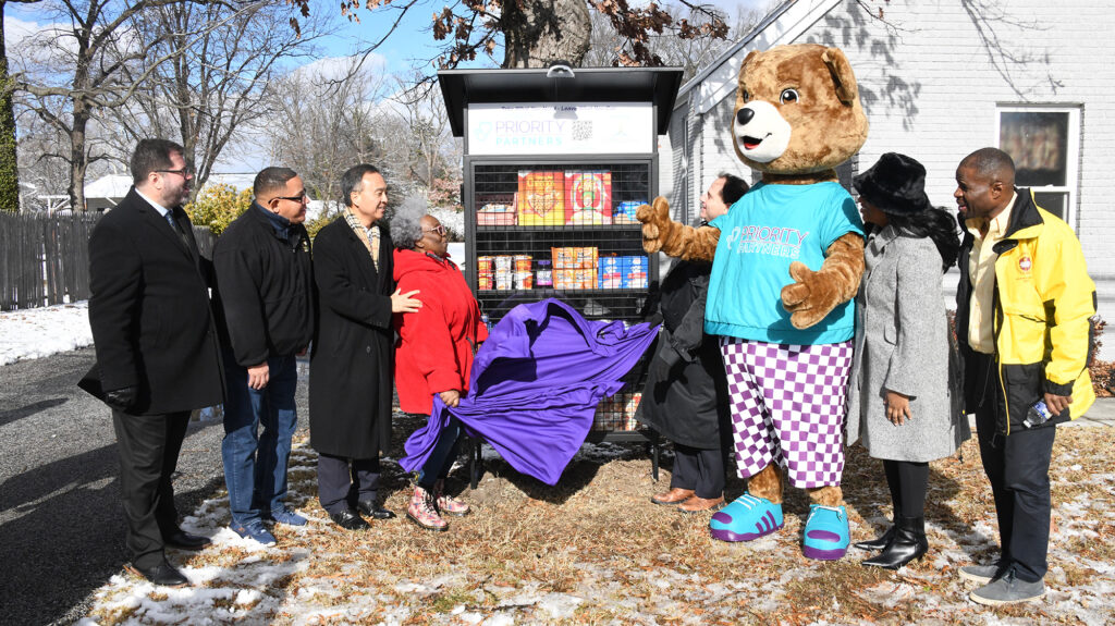 A food pantry cupboard is unveiled as several people and a mascot watch.