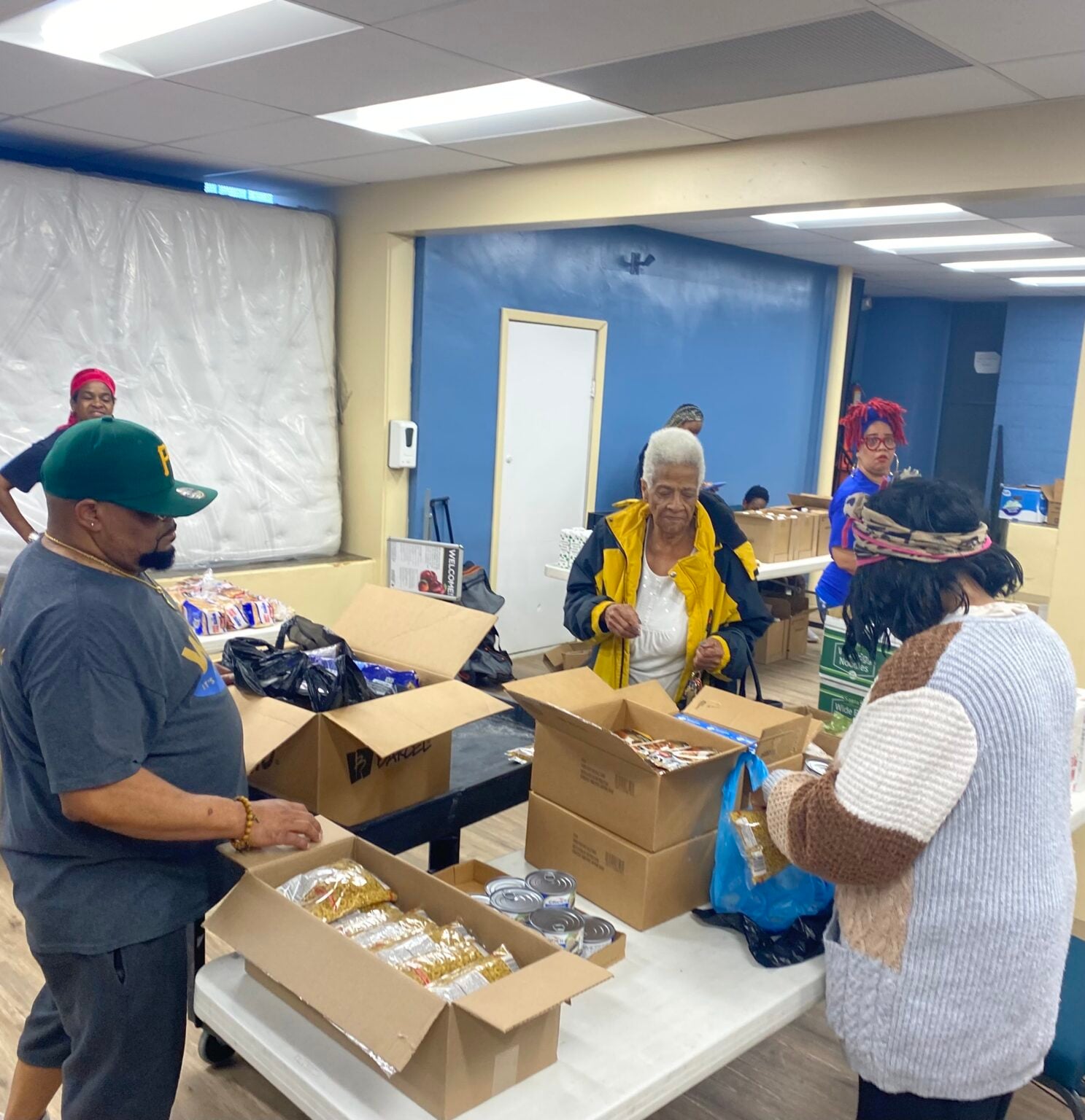 People standing at looking at boxes sitting on tables that have food items.