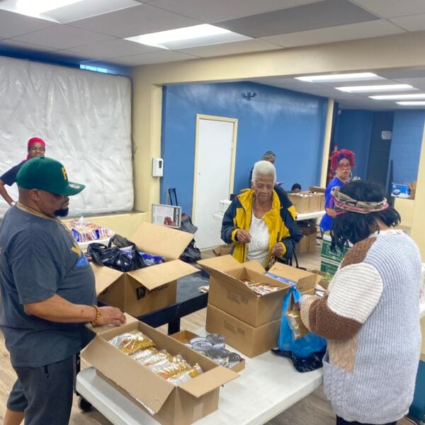 People standing at looking at boxes sitting on tables that have food items.