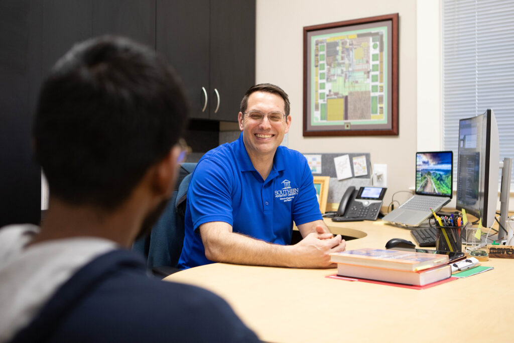 A male college professor speaking with a male college student in his office.