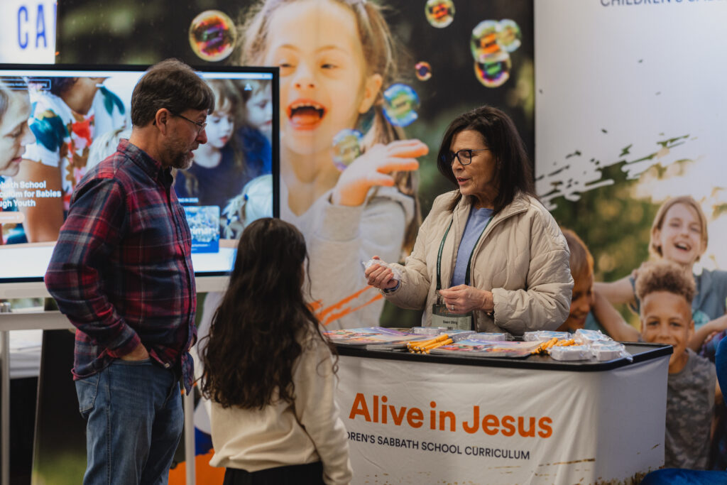 Adult male and adolescent girl listen to adult woman at an exhibit booth focused on children.