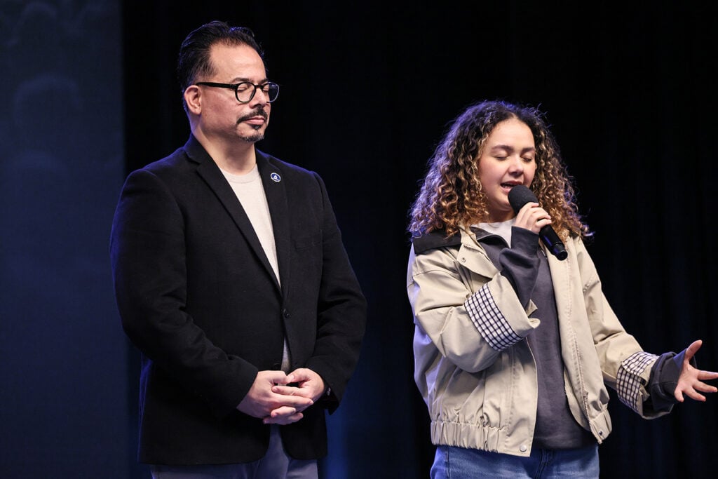 A man and young woman stand together as she prays on a stage. 