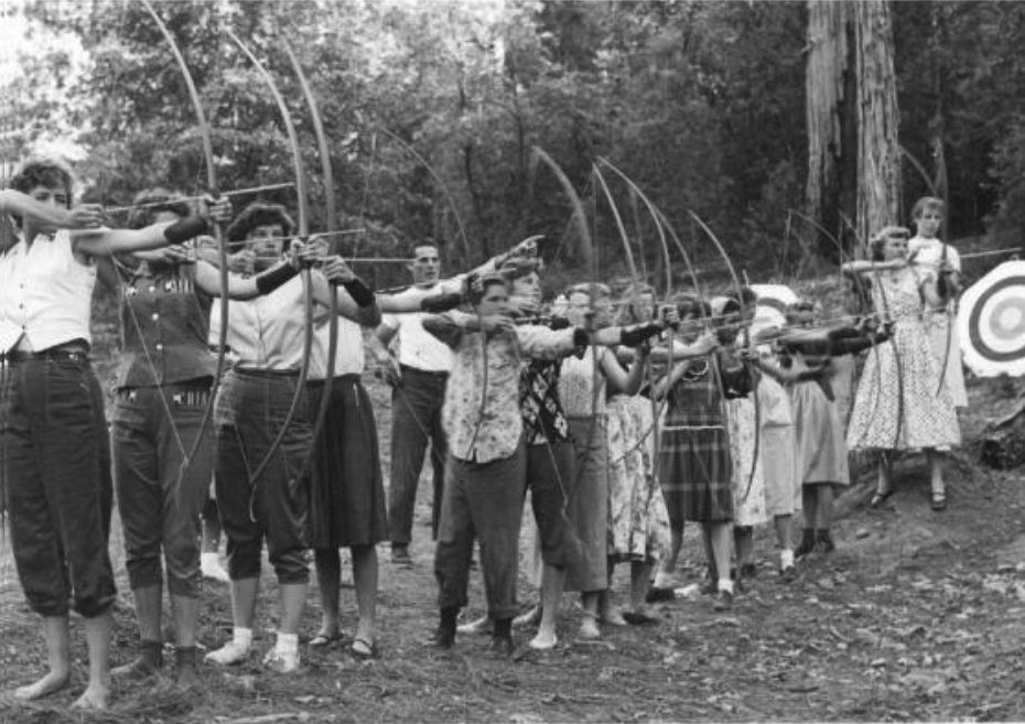 Black and white photo of young women in a line about to shoot an arrow in a group archery class.