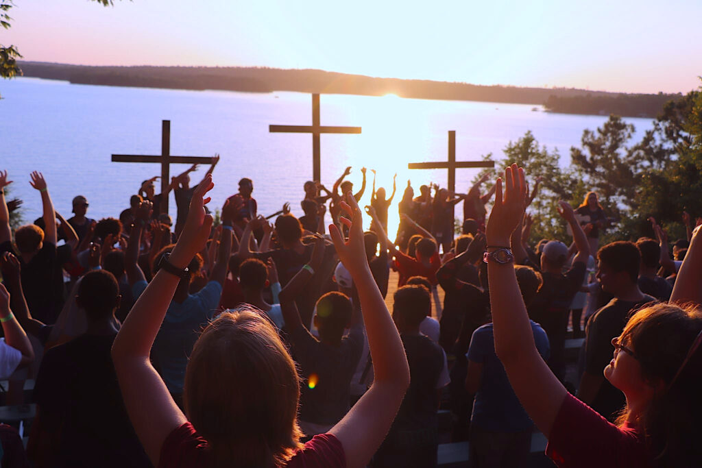 Campers worship outside, arms upraised, looking toward three crosses on a makeshift stage.