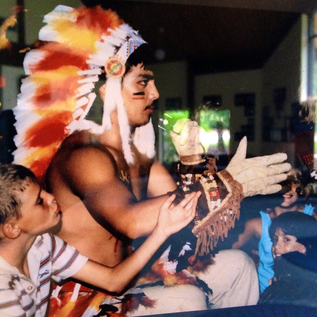 A young man in a Native headdress embraced by a young boy, as other children look on.