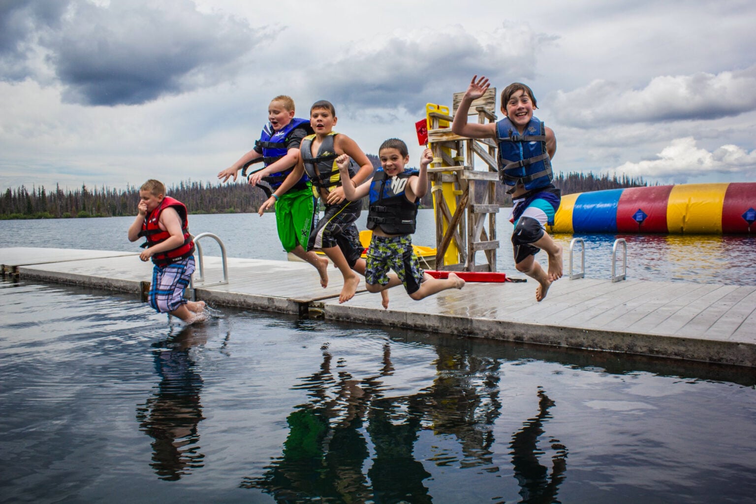Several boys jump off a deck at a lake.