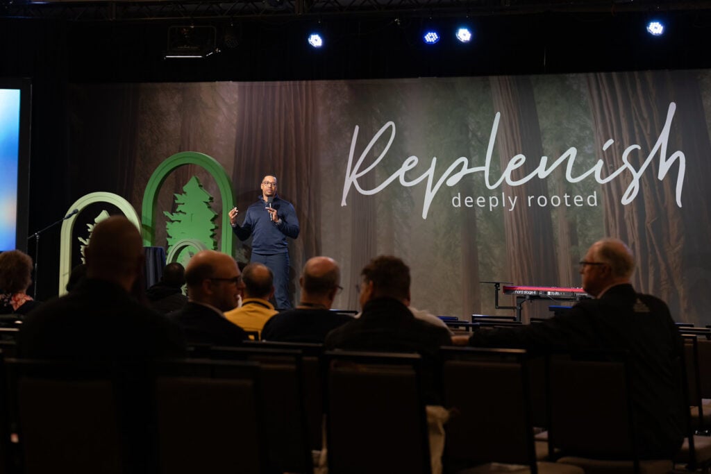 Wide shot of a speaker onstage with the word “Replenish” displayed behind him as attendees listen.