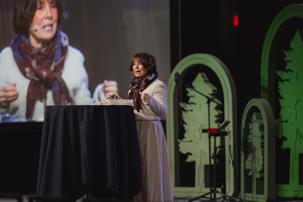Women speaks on stage behind a small table while her image appears on a large screen behind her.