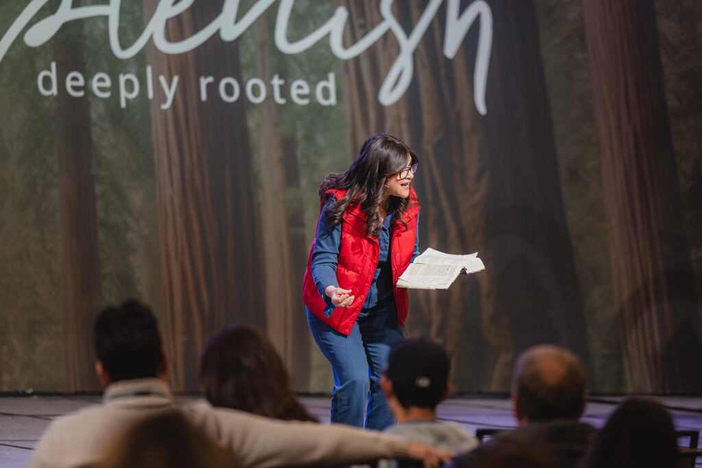 A woman speaks on stage holding a Bible and leaning toward the audience. 