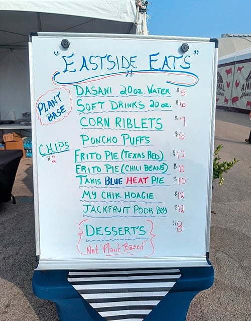 The image shows a whiteboard menu displayed on a blue stand at  an outdoor food booth.