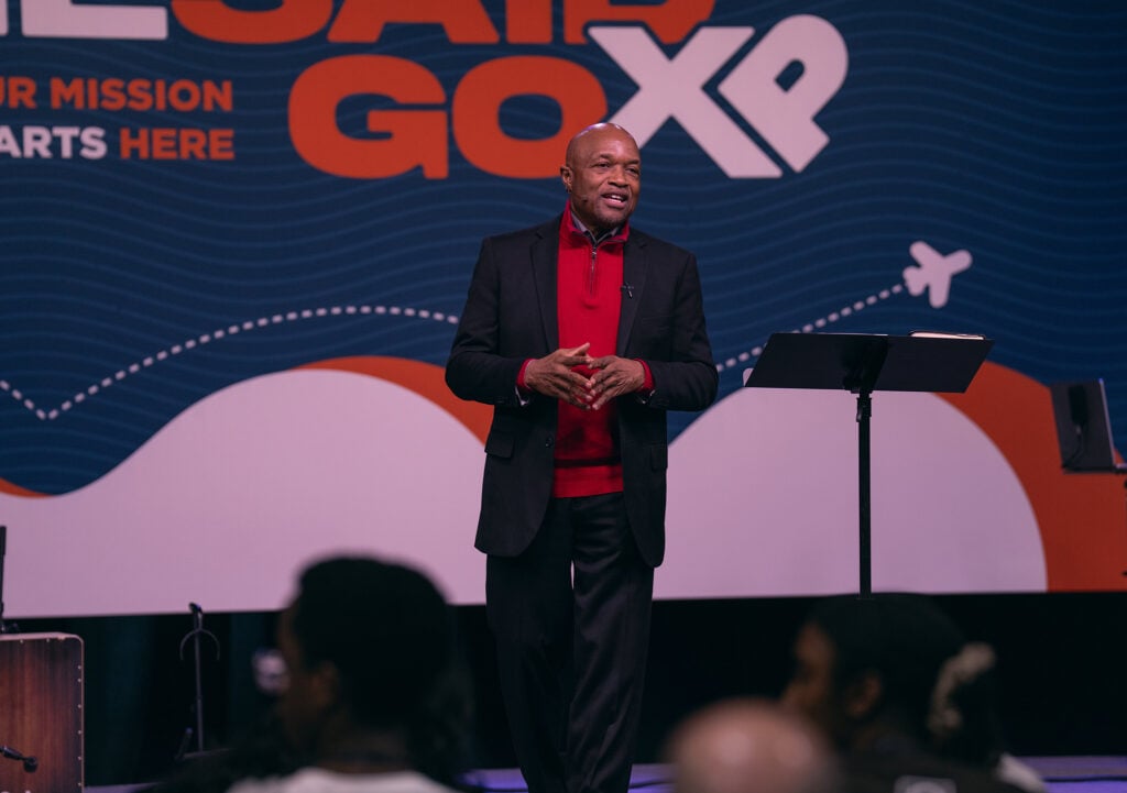 A man stands on stage in front of a screen reading He Said Go XP.