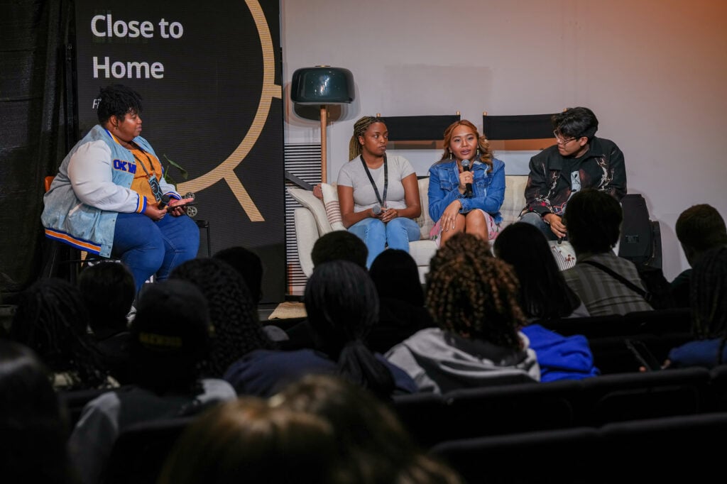 A panel with a facilitator on a separate chair and three panelists on a couch. Heads of audience in foreground.