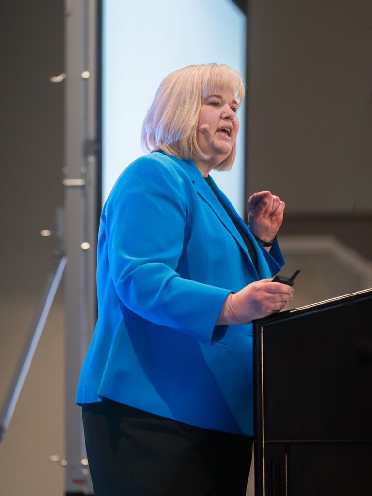 A woman in a blue suit stands at a podium speaking