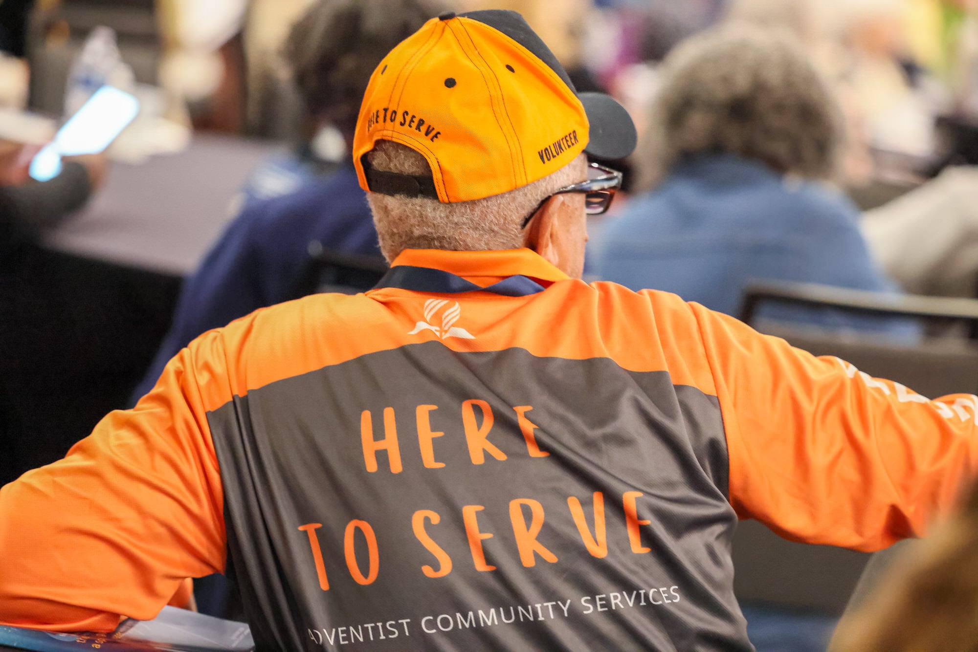 An Adventist Community Services volunteer sitting at a meeting. Photo shows the Here to Serve logo on the back of the shirt and of his hat.
