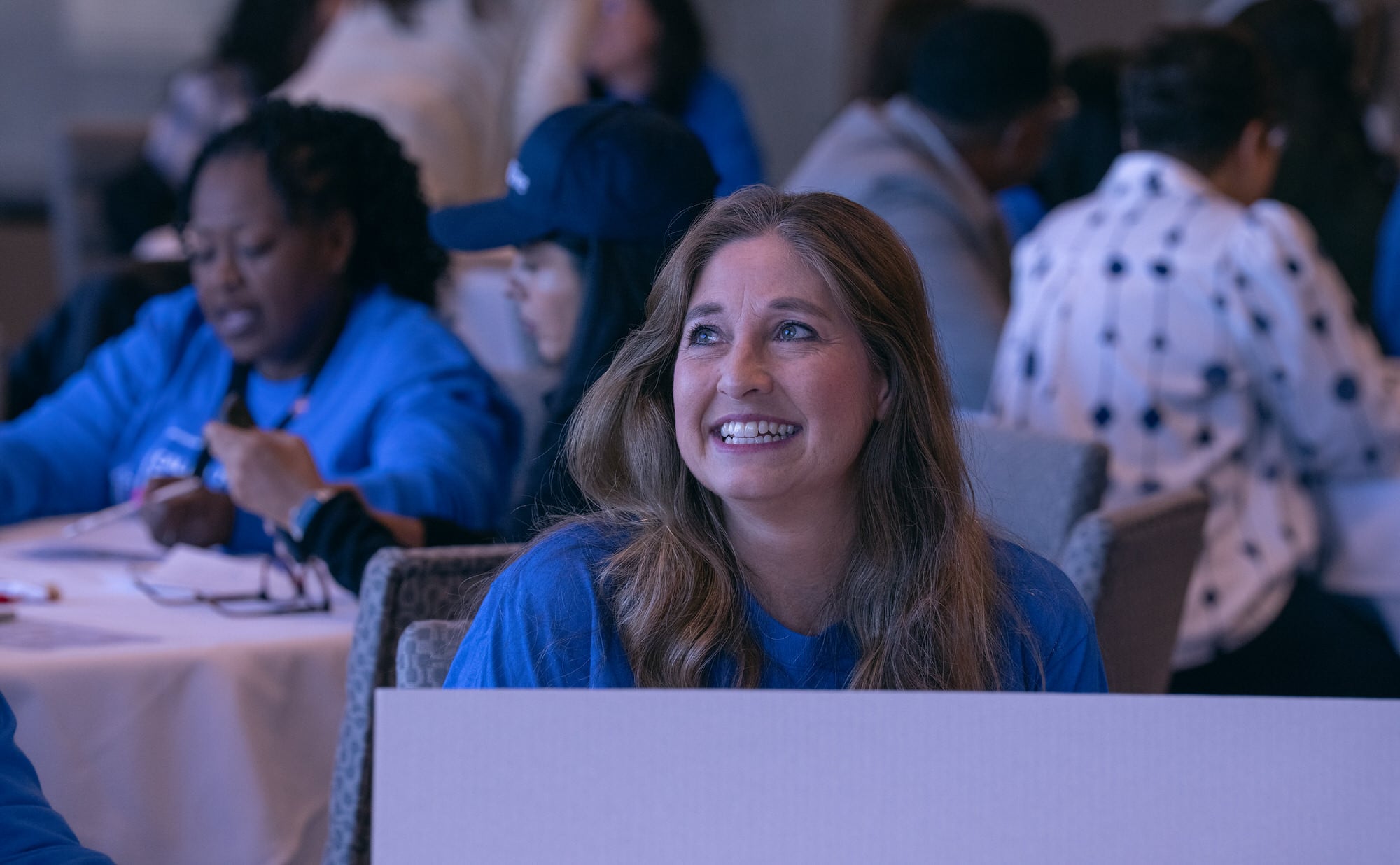 Smiling blond haired woman at a conference table looks up towards the stage.