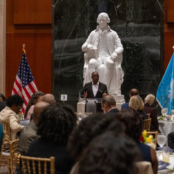 A man stands in front of a crowd speaking with a prominent white statue behind him.
