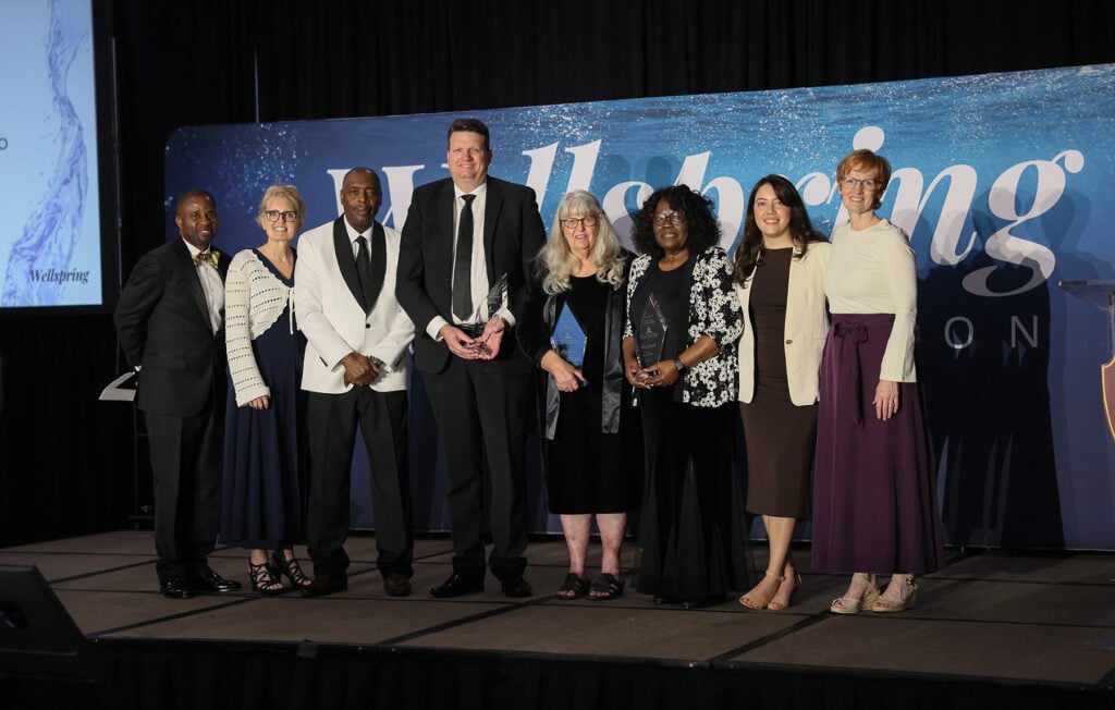 A group of men and women, some holding awards, stand on the Wellspring stage.