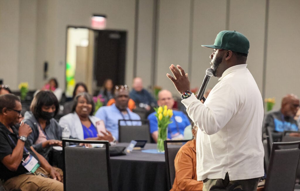 A man in a baseball cap speaks in front of an audience