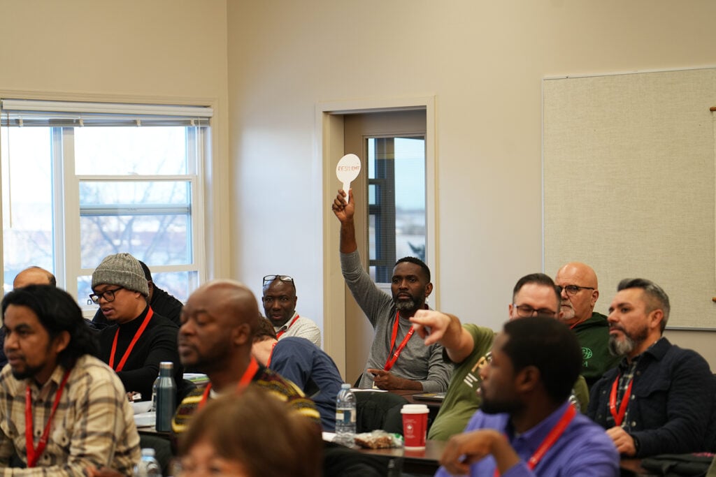 A man in a classroom setting surrounded by other menholds up a cardboard paddle reading "Resilience."