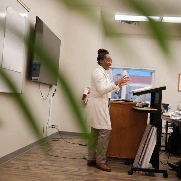 A woman stands at a podium speaking, with photo framed by green leaves