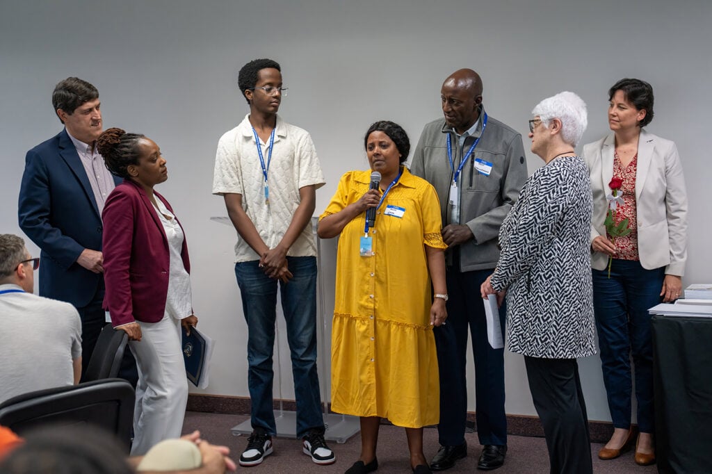 A woman speaks into a microphone while surrounded by her family and church leaders during a ceremony recognizing her years of missionary service.