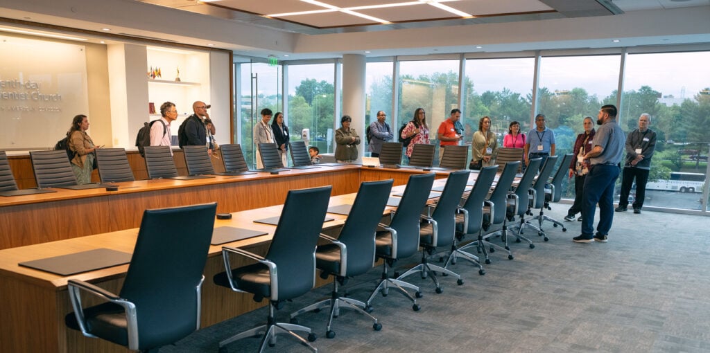 Several people stand in a boardroom around a long table. 