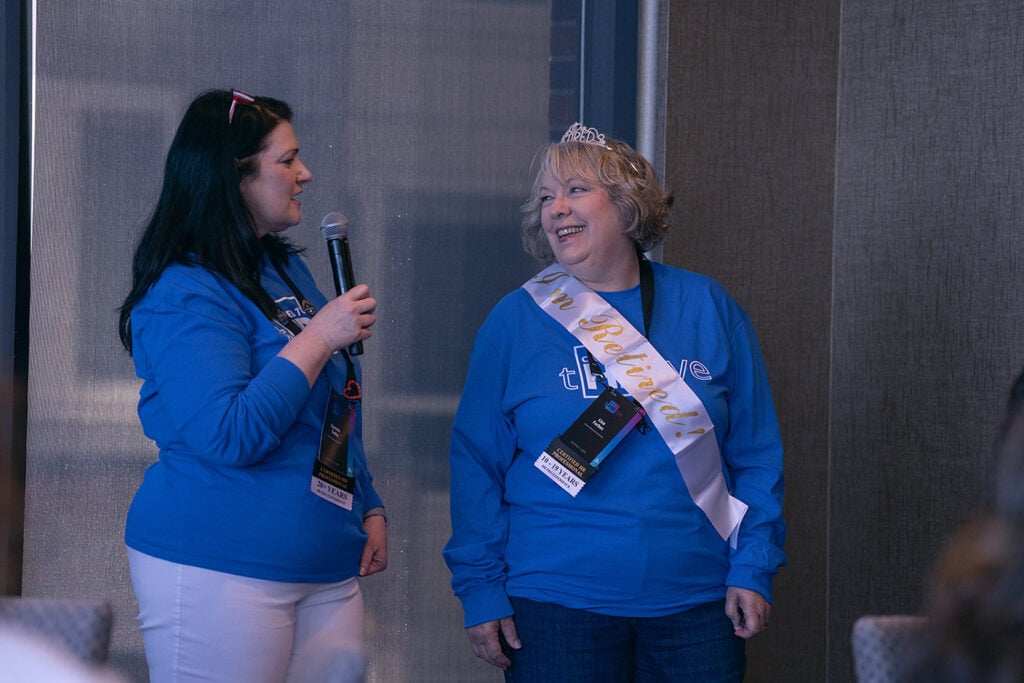 One woman stands with a mic, speaking, as another, wearing a tiara and retirement sash, smiles in her direction