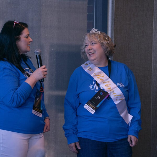 One woman stands with a mic, speaking, as another, wearing a tiara and retirement sash, smiles in her direction
