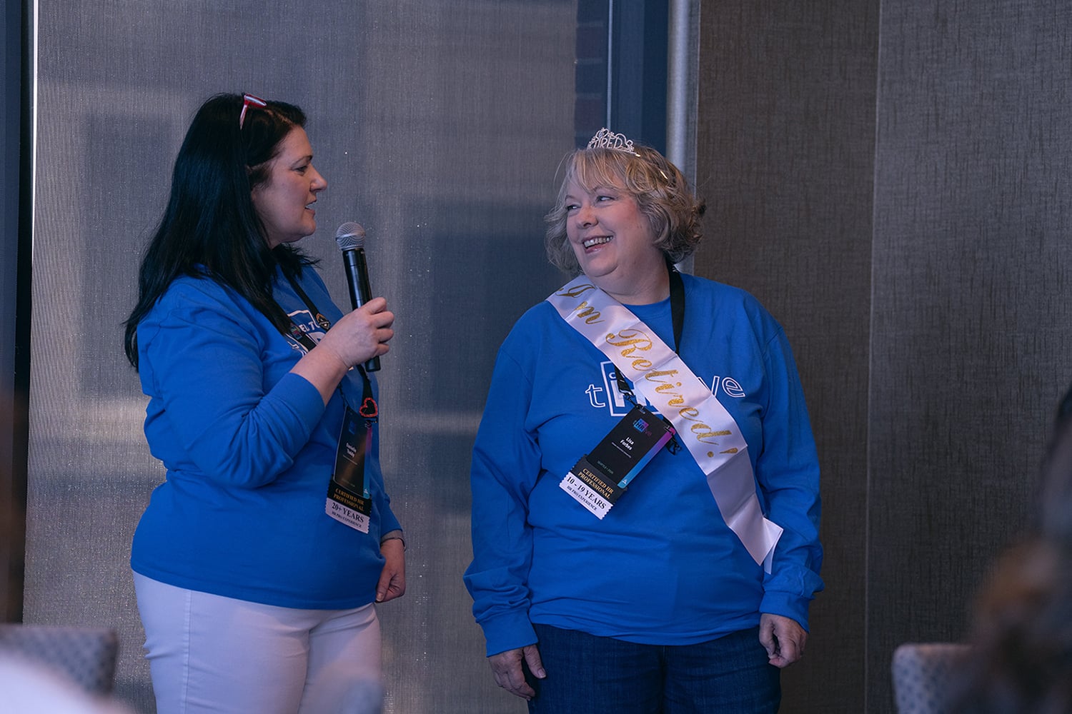 One woman stands with a mic, speaking, as another, wearing a tiara and retirement sash, smiles in her direction