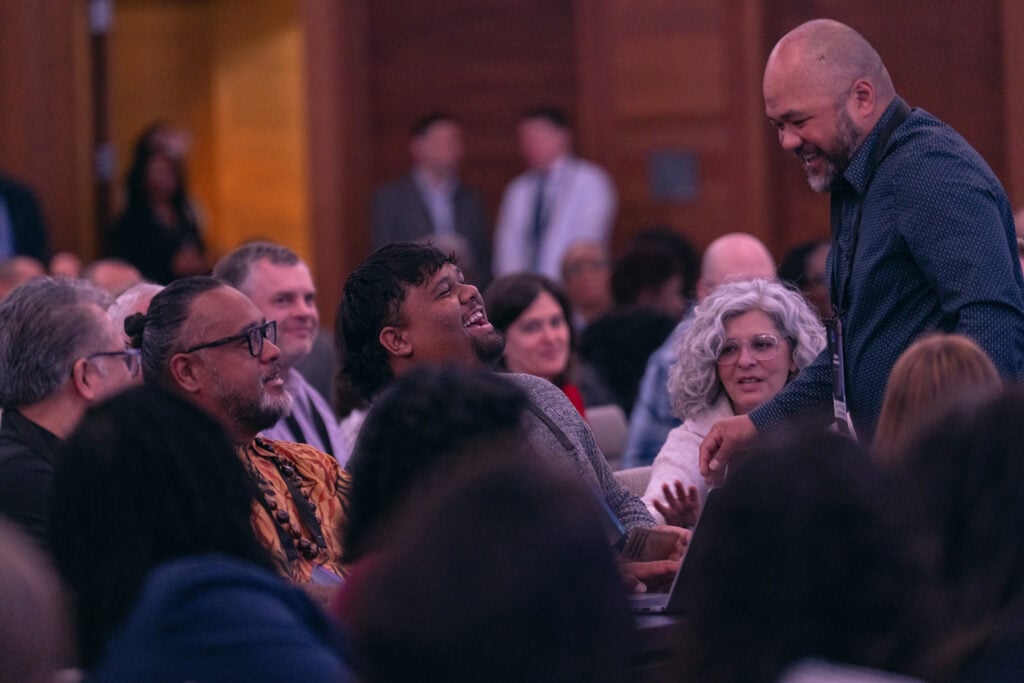 A man stands up speaking to two man who are seated in a crowded conference room. All are laughing.