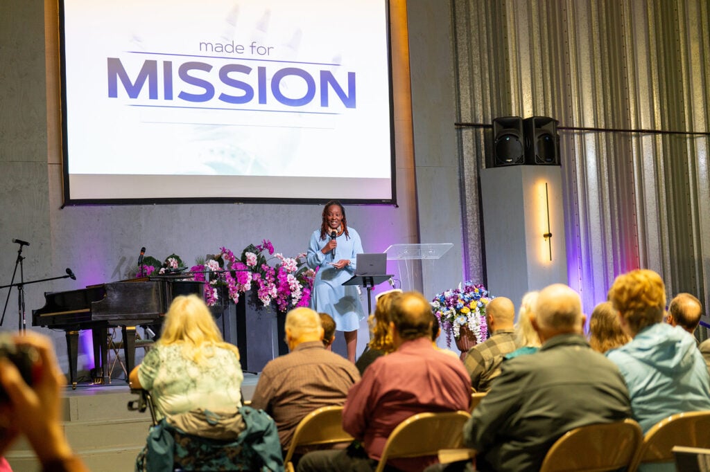 Orna Garnett speaks onstage at camp meeting in front of a presentation slide reading “Made for Mission.”