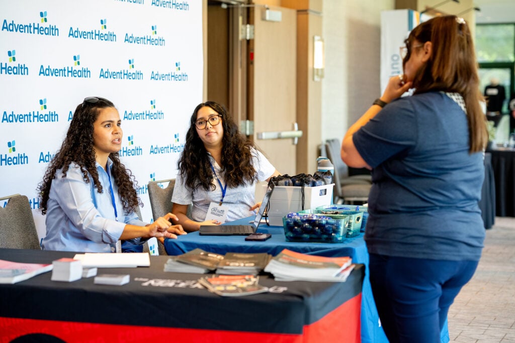 An SAC attendee speaks with two AdventHealth representatives in the exhibit hall.