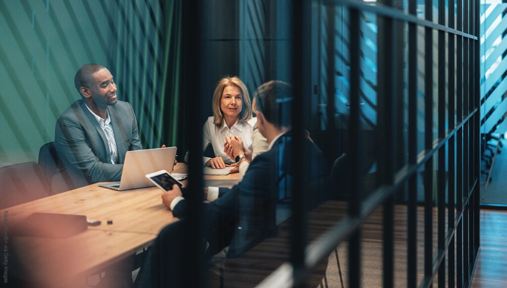 Business professionals sit together in a contemporary glass-walled office, engaged in a focused consultation. Reviewing information on a laptop and mobile device, they discuss strategies, insights, and solutions for business growth. The scene captures the essence of small business consulting, teamwork, and data-driven decision-making in a dynamic corporate environment