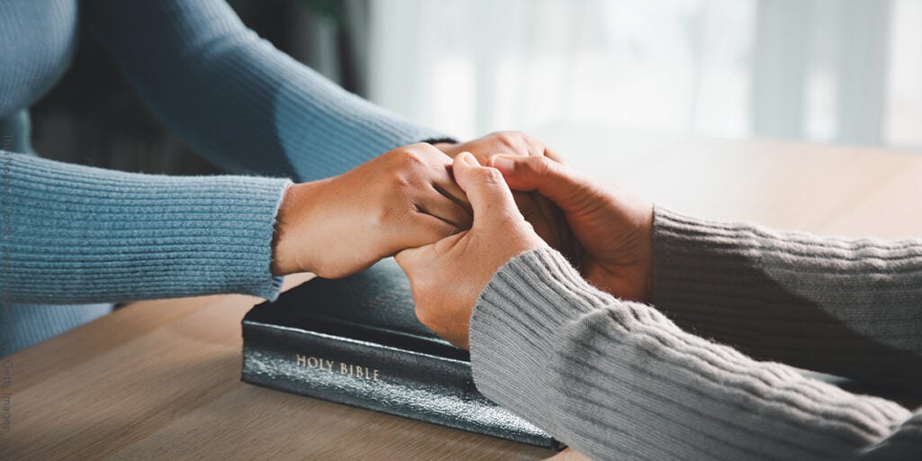 Close-up of religious people holding hands in prayer over the Holy Bible on a wooden table, offering comfort, spiritual support, hope, and Christian faith together in a bright room.