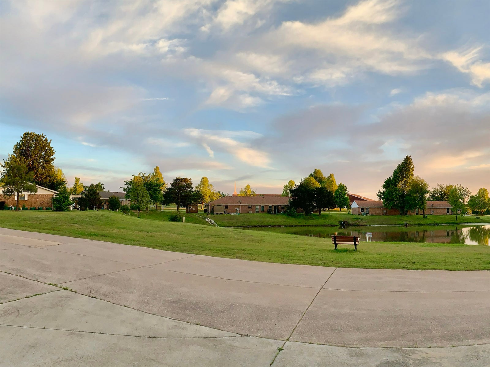 Roadside view of a pond with homes and apartments beyond at Summit Ridge Retirement Village during early evening light