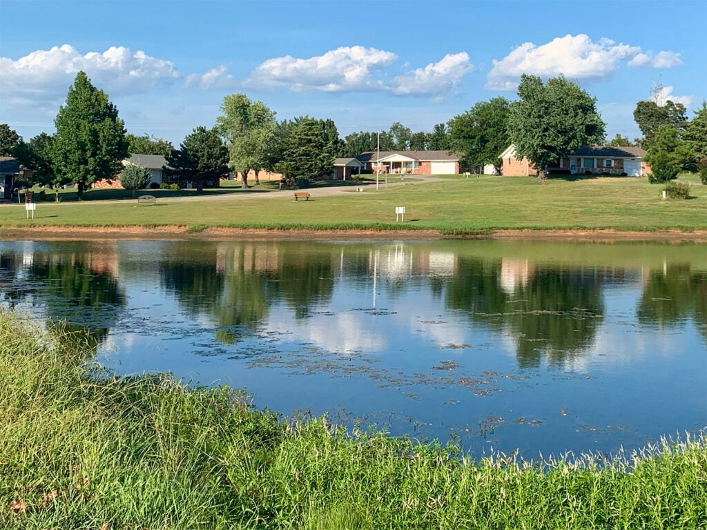 Pond with grassy foreground, benches, and homes surrounded by trees under a blue summer sky