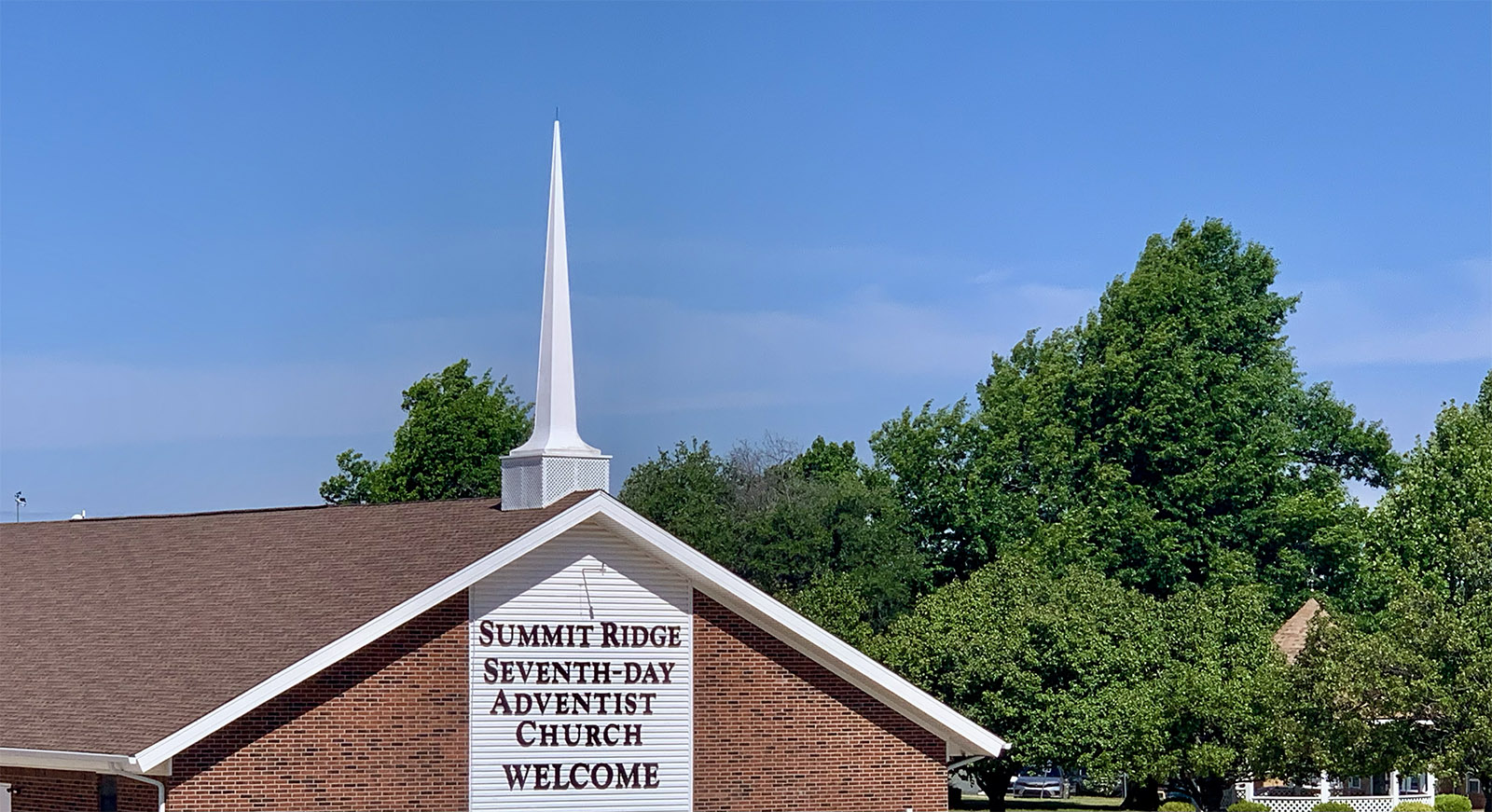 Brick church with white steeple and nearby gazebo surrounded by large green trees beneath a blue sky with clouds