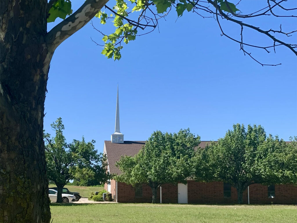 Side view of a brick church with white steeple framed by tree branches and green leaves