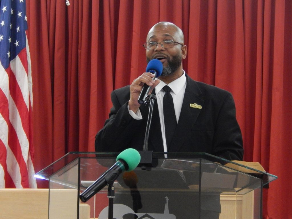 A man in a dark suit singing into a microphone at the pulpit, with an American flag and red curtain behind him