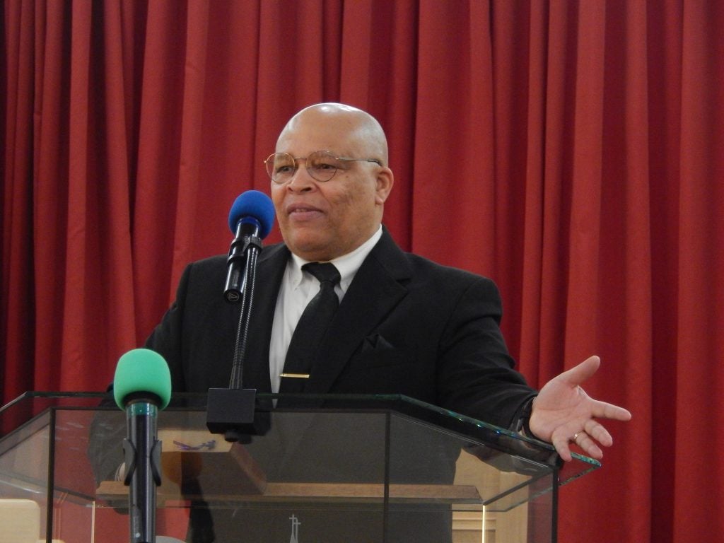 A man in a dark suit speaking at the pulpit with one hand open, in front of a red curtain