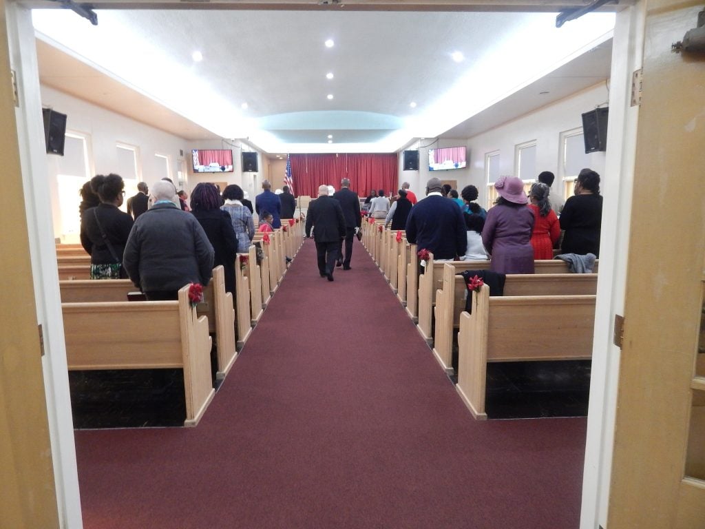 Church members standing among the wooden pews in the Temple of Praise sanctuary