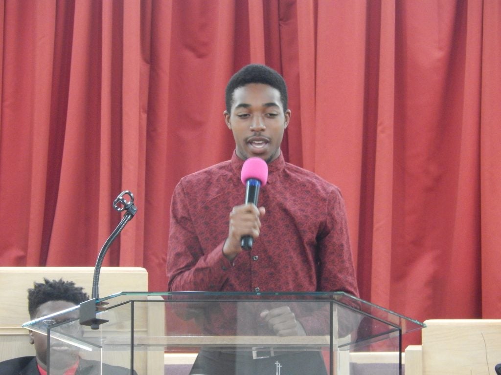 A young man in a maroon shirt speaking into a microphone at the pulpit